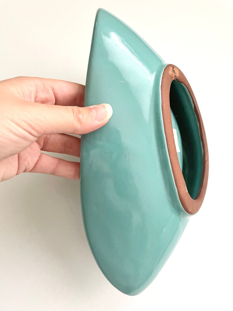 May include: A light blue ceramic planter with a rounded, oval shape and a brown rim. The planter is designed to be hung on a wall.