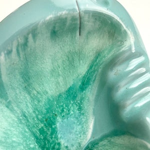 May include: A light blue ceramic ashtray with a green and white marbled glaze. The ashtray has a deep, rounded bowl with a slightly raised rim.