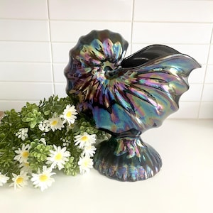 May include: Iridescent ceramic nautilus shell vase. The vase has a dark, glossy finish with a rainbow sheen. The vase is on a white surface next to a bundle of greenery and white flowers.