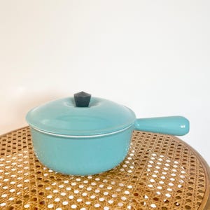 May include: A turquoise enamel saucepan with a lid and a single handle. The lid has a dark grey knob. The pan is on a woven wicker surface. The design is simple and classic. The pan is approximately 15 cm in diameter.