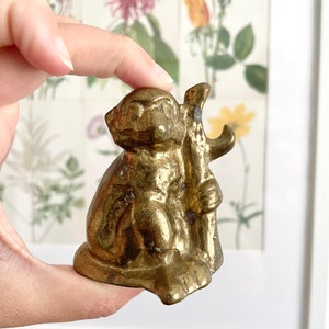 May include: A small, gold-coloured metal monkey figurine holding a stick. The monkey is sitting with its legs crossed and its head tilted to the side.