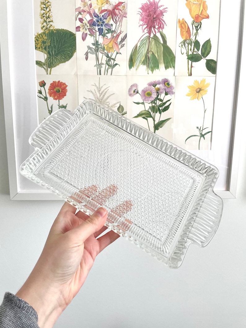 Puede incluir: Una bandeja rectangular de vidrio transparente con un patr&oacute;n de diamantes y asas acanaladas. La bandeja se sostiene contra un fondo de estampados bot&aacute;nicos enmarcados con flores coloridas. Una pieza decorativa para servir.