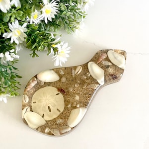 May include: A brown resin spoon rest with a sand and seashell design. The spoon rest is shaped like a fish and has a variety of seashells embedded in the resin.