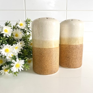 May include: A pair of ceramic salt and pepper shakers with a white, yellow and brown speckled glaze. The shakers are round in shape and have a small opening on top for dispensing salt and pepper.