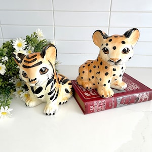 May include: Two ceramic animal figurines. One is a tiger cub with black stripes on a cream base. The other is a leopard cub with black spots on a cream base. Both have large eyes and are sitting down.