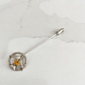 Puede incluir: Broche de plata con forma de cardo con un centro de strass ámbar. La broche está unida a un largo alfiler de plata.