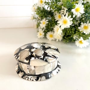 May include: A decorative, round jewelry box with a lid. The box is made of a mosaic of black, white, and gray pieces. The box sits on a white surface, with a bouquet of white daisies in the background.