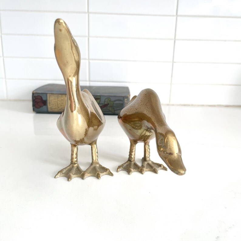 May include: Two brass duck figurines. One duck stands upright with its head raised, while the other bends down. The ducks have detailed feather and foot designs. The background includes a decorative box and a white tiled wall.