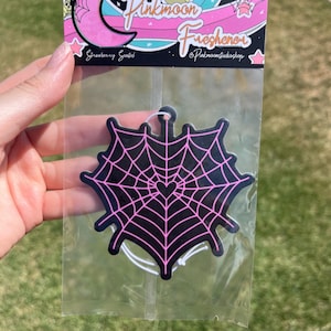 May include: Pink car air freshener with a black and pink spiderweb design. The air freshener is packaged in a clear plastic bag with a white string.