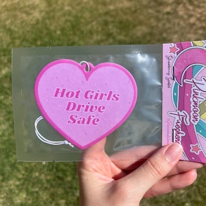 Puede incluir: Ambientador de coche en forma de corazón rosa con el texto "Hot Girls Drive Safe" en letras blancas. El ambientador está empaquetado en una bolsa de plástico transparente.