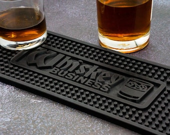 Puede incluir: Tapete de barra negro con el texto "Whiskey Business" y una imagen estilizada de un vaso de whisky. La tapete tiene una superficie texturizada con pequeños puntos en relieve.