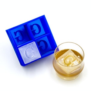 May include: Blue ice cube tray with four compartments, each with the letter "G" molded into the bottom. A clear glass with whiskey and a single ice cube with the letter "G" is next to the tray.