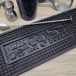Może przedstawiać: Czarna mata barowa z napisem "WIN OR LOSE WE STILL BOOZE" i grafiką sportową. Zawiera jigger, łyżkę do mieszania, sitko do koktajli i nakrapiany kubek. Mata wykonana jest z teksturowanego materiału gumowego.