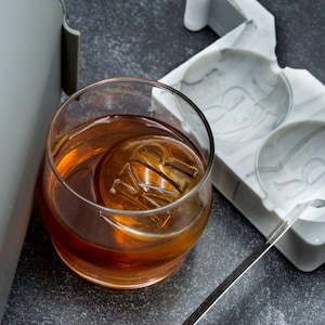 May include: A rocks glass filled with amber liquid and a large ice sphere with an embossed design. A white and gray ice mold and a metal ice tool are also visible. The drink is on a dark gray surface.