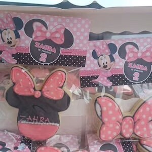 K&ouml;nnte beinhalten: Ein Set mit pinkfarbenen und schwarzen Keksen im Minnie-Mouse-Design, verziert mit rosa Punkten und dem Namen "Zahra" in rosa Schrift. Die Kekse sind in durchsichtigen Cellophanbeuteln verpackt.