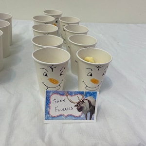 May include: A table with white paper cups decorated with snowman faces. The cups are filled with yellow snacks. A sign with a cartoon reindeer and the text "Snow Flurries" is in the foreground.