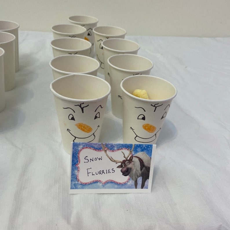 May include: A table with white paper cups decorated with snowman faces. The cups are filled with yellow snacks. A sign with a cartoon reindeer and the text "Snow Flurries" is in the foreground.