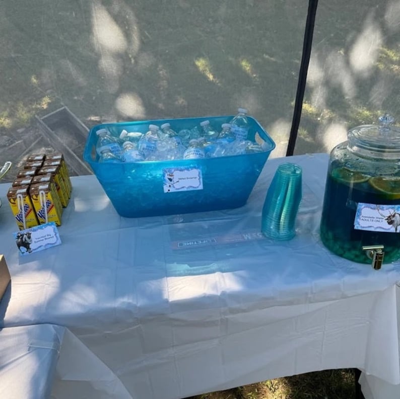 May include: A white table covered with a white tablecloth with a blue plastic tub filled with water bottles and ice. There is a glass pitcher filled with blue liquid and fruit. There are also small boxes of chocolate milk and blue plastic cups.