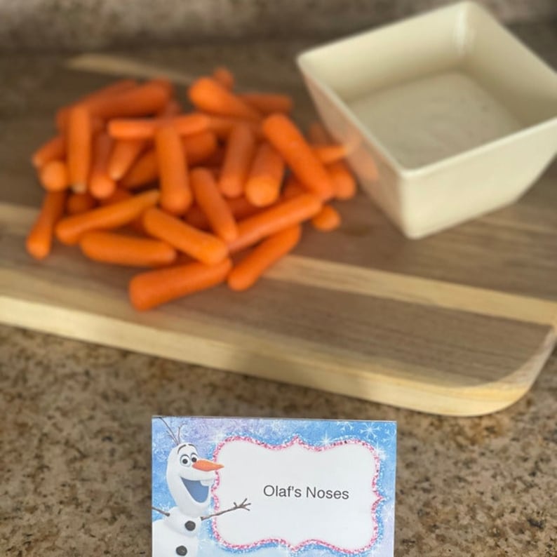 May include: A wooden cutting board with a small white bowl filled with a white sauce. A pile of orange baby carrots are on the board. A blue and pink card with a cartoon Olaf from Frozen says "Olaf's Noses".
