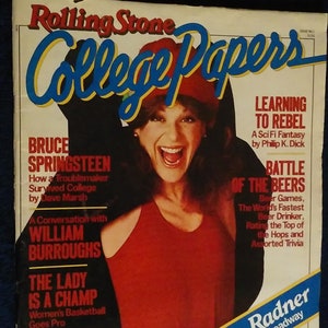 May include: A magazine cover for Rolling Stone College Papers featuring Gilda Radner, a woman with red hair wearing a red tank top and a red hat, smiling with her mouth open. The cover also features articles about Bruce Springsteen, William Burroughs, and a battle of the beers.