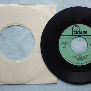 Puede incluir: Un disco de vinilo negro con una etiqueta blanca. La etiqueta tiene el texto "Fontana" en un círculo, y el título de la canción "You've Got To Hide Your Love Away" de "The Silkie".