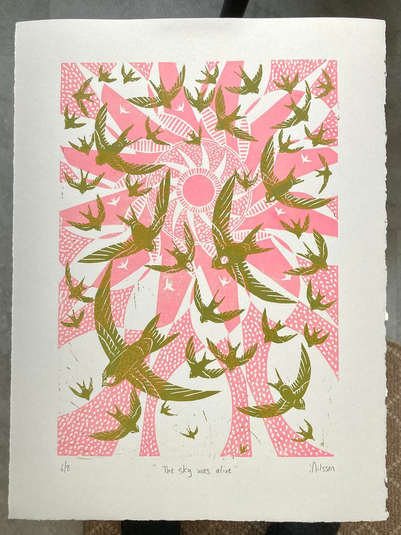 May include: A print featuring a flock of birds in flight, rendered in shades of olive green and pink. The birds are set against a stylized sunburst design. The text "The sky was alive" is at the bottom.