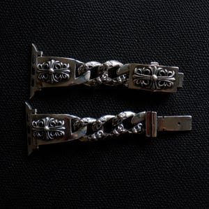 May include: Two silver-colored watch bands with a chain link design. Each band features ornate cross-like embellishments on the ends and clasp. The bands are displayed on a dark, textured surface.