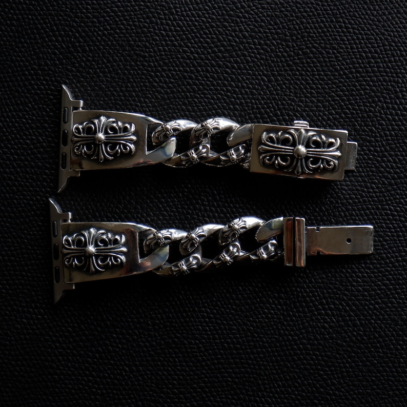 May include: Two silver-colored watch bands with a chain link design. Each band features ornate cross-like embellishments on the ends and clasp. The bands are displayed on a dark, textured surface.