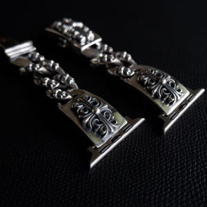 May include: Two silver watch bands with ornate cross designs. The bands are made of linked metal pieces and feature a decorative clasp. The watch bands are displayed on a dark surface, highlighting the intricate details of the metalwork.