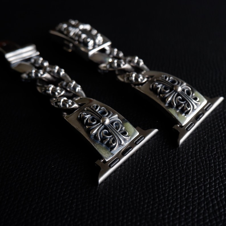 May include: Two silver watch bands with ornate cross designs. The bands are made of linked metal pieces and feature a decorative clasp. The watch bands are displayed on a dark surface, highlighting the intricate details of the metalwork.