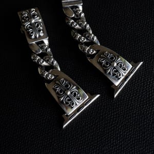 May include: Close-up of two silver watch band links with ornate, symmetrical floral and cross designs. The links are connected by a chain of similar design. The watch band is on a dark background.