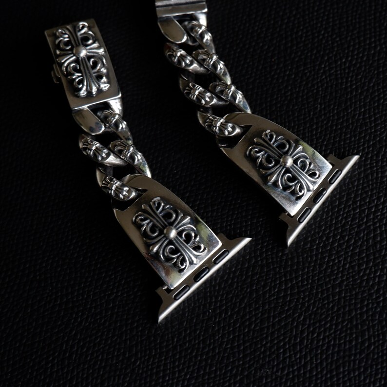 May include: Close-up of two silver watch band links with ornate, symmetrical floral and cross designs. The links are connected by a chain of similar design. The watch band is on a dark background.