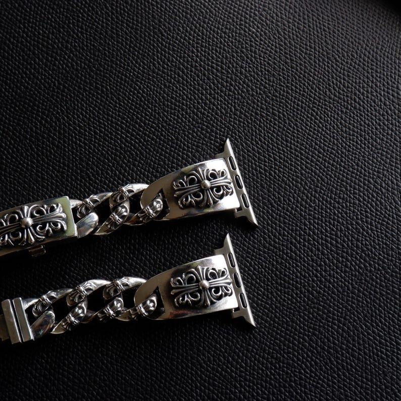 May include: Two silver watch bands with ornate, symmetrical designs. The bands feature a chain-link style and decorative clasps. The watch bands are displayed against a dark, textured background.