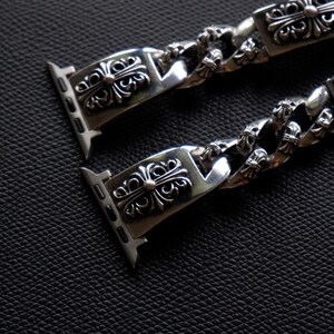 May include: Silver-toned watch band with a detailed, ornate design. The band features a series of decorative rectangular links with a floral cross motif, connected by a chain-like structure. The watch band is shown against a dark, textured background.