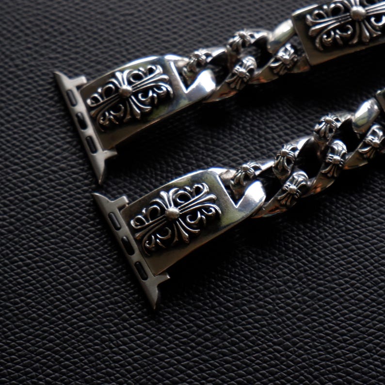 May include: Silver-toned watch band with a detailed, ornate design. The band features a series of decorative rectangular links with a floral cross motif, connected by a chain-like structure. The watch band is shown against a dark, textured background.