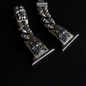 May include: A pair of silver-colored watch bands with ornate, cross-shaped designs. The bands feature a chain-link style and are designed to attach to a smartwatch. The bands are displayed against a dark background.