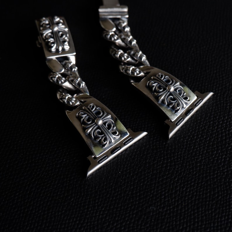 May include: A pair of silver-colored watch bands with ornate, cross-shaped designs. The bands feature a chain-link style and are designed to attach to a smartwatch. The bands are displayed against a dark background.