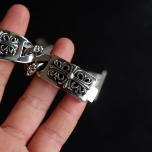 May include: A silver bracelet with a decorative cross design. The bracelet is made of a shiny metal and features intricate detailing on each link. The clasp is rectangular with a cross design and a small silver ball in the center.