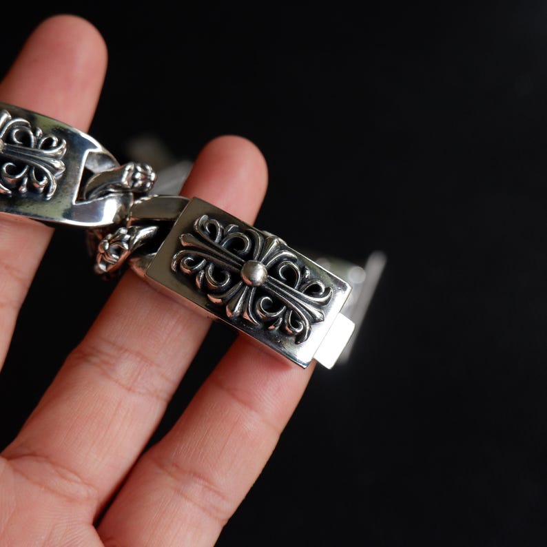 May include: A silver bracelet with a decorative cross design. The bracelet is made of a shiny metal and features intricate detailing on each link. The clasp is rectangular with a cross design and a small silver ball in the center.