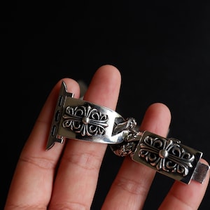 May include: A silver-colored watch band with an ornate cross design. The band is made of metal and features a series of decorative cross-shaped links. The watch band is designed to attach to a smartwatch.