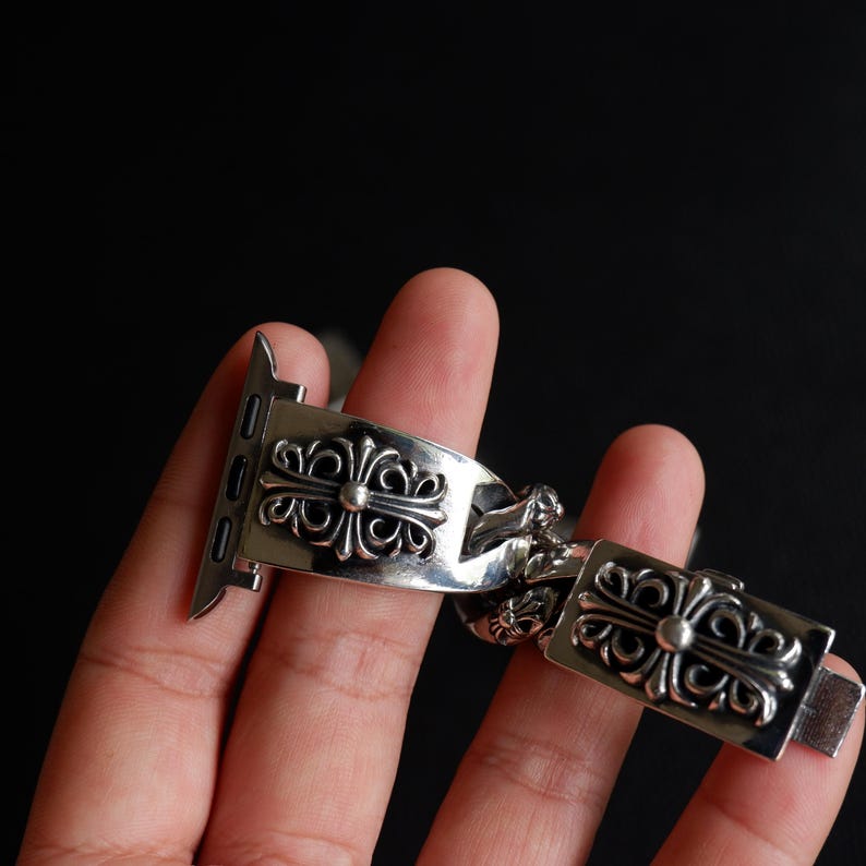 May include: A silver-colored watch band with an ornate cross design. The band is made of metal and features a series of decorative cross-shaped links. The watch band is designed to attach to a smartwatch.
