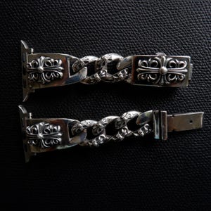 May include: A silver-colored watch band with a chain link design. The band features decorative cross-shaped embellishments and a clasp. The watch band is shown against a black background.
