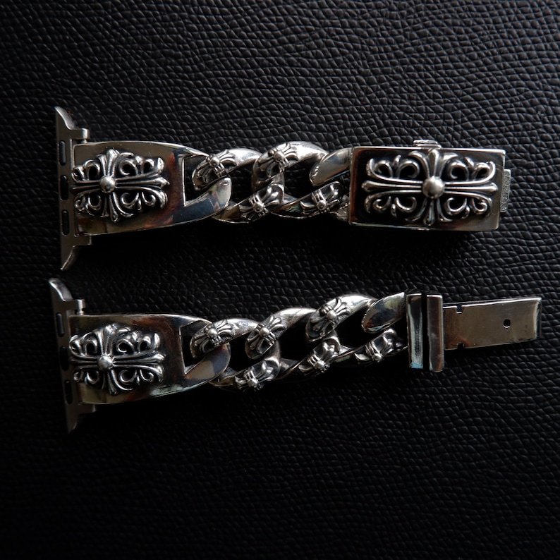 May include: A silver-colored watch band with a chain link design. The band features decorative cross-shaped embellishments and a clasp. The watch band is shown against a black background.