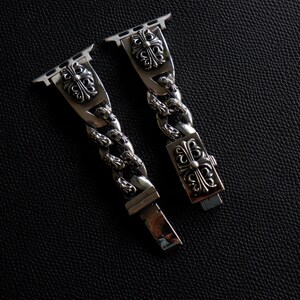 May include: Silver-toned watch band with a chain link design. The band features decorative cross-like emblems on the connectors and clasp. The watch band is shown against a dark, textured background.