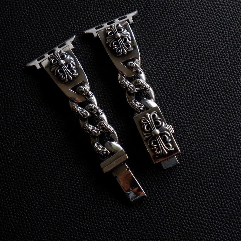 May include: Silver-toned watch band with a chain link design. The band features decorative cross-like emblems on the connectors and clasp. The watch band is shown against a dark, textured background.