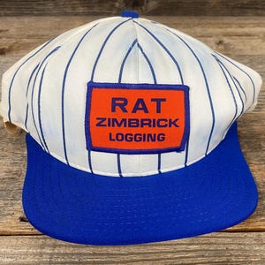 May include: A white baseball cap with blue vertical stripes and a blue brim. The cap features a rectangular patch with the words "RAT ZIMBRICK LOGGING" in blue text on an orange background.