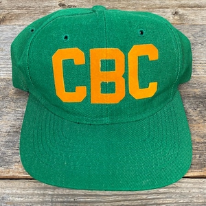 Può includere: Un cappellino da baseball verde con visiera piatta. Il cappellino presenta le lettere arancioni "CBC" sul davanti. Il cappello è realizzato in un tessuto strutturato e ha un design classico e strutturato.