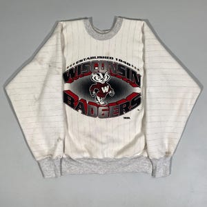 May include: White and gray striped sweatshirt featuring the Wisconsin Badgers logo. The logo includes a badger mascot and the words "WISCONSIN BADGERS" in red and black. The sweatshirt has a gray collar and cuffs.