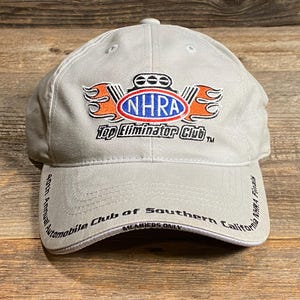 May include: A light grey baseball cap with the NHRA Top Eliminator Club logo in orange, white, and blue. The cap also displays text that reads "40th Annual Automobile Club of Southern California NHRA Finals".