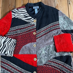 May include: A long-sleeve jacket featuring a patchwork design with black, red, grey, and white zebra print panels. The garment has a mandarin collar, four circular textured buttons, and a black interior lining with an Indigo Moon brand label.